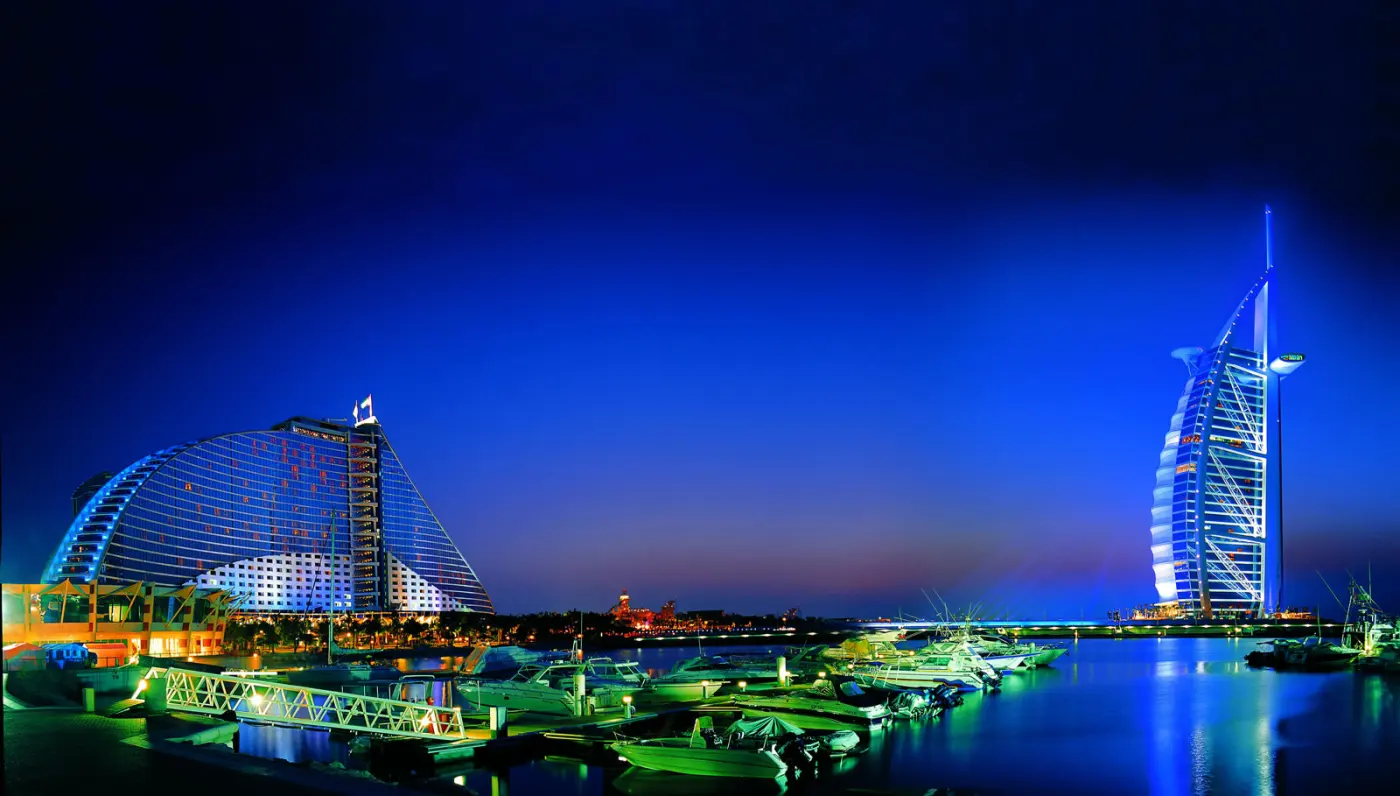Ночная панорама пляжной Джумейры: подсвеченный отель-волна Jumeirah Beach Hotel слева и отель-парус Бурдж-эль-Араб справа, между ними катера у причала